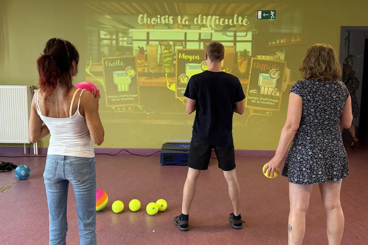 Trois adultes debout face au mur interactif, balles en main, devant un jeu de recettes projeté