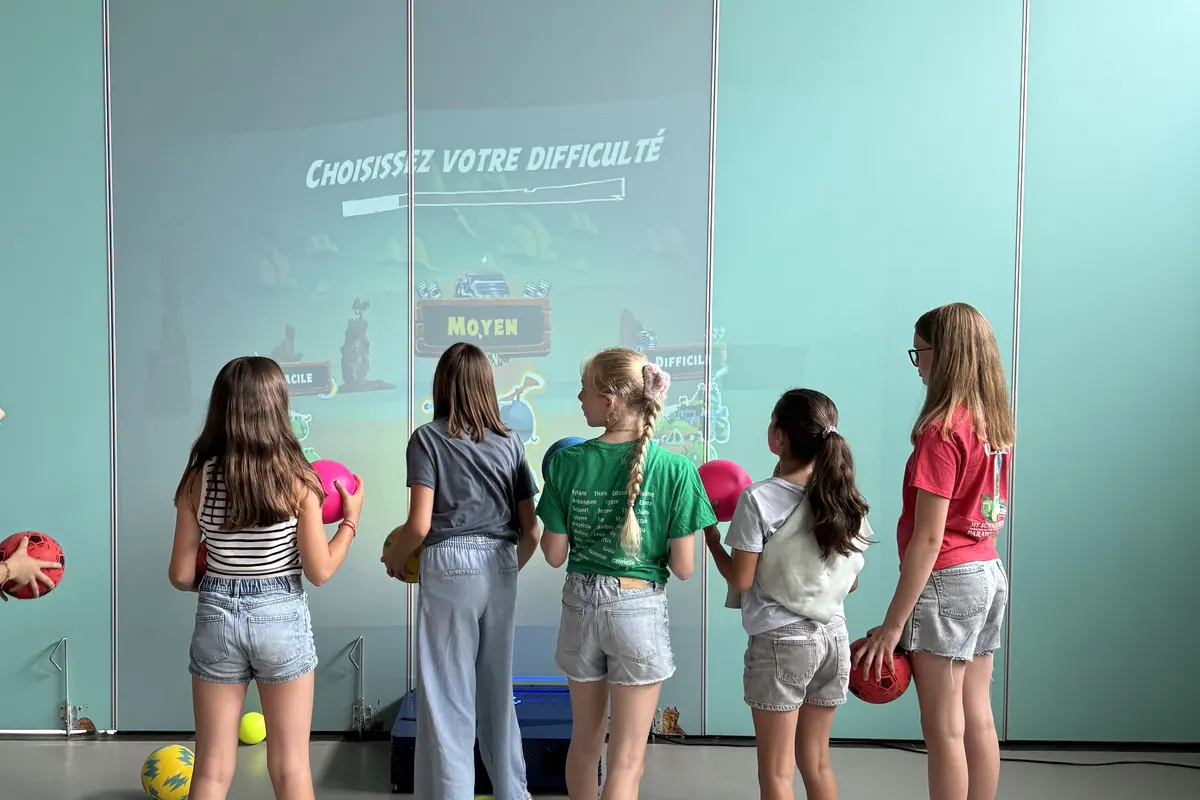 Élèves face au mur interactif choisissant leur niveau de difficulté, balles en main, dans la salle de gym de leur école