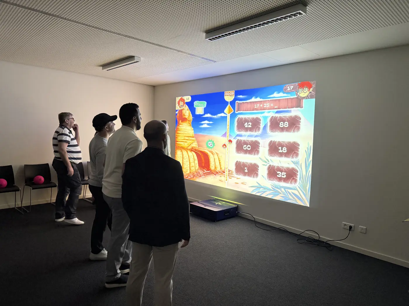 Un jeu de calcul projeté sur le mur blanc d'une salle de réunion, le boîtier posé au sol, quatre collaborateurs debout face à la projection