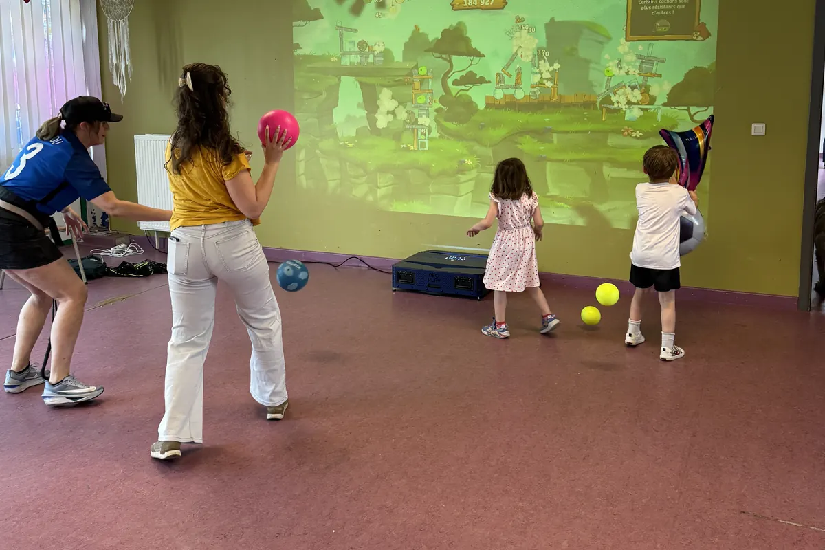 Deux adultes et deux jeunes enfants, balles en main, face à un jeu projeté sur le mur d'une salle polyvalente